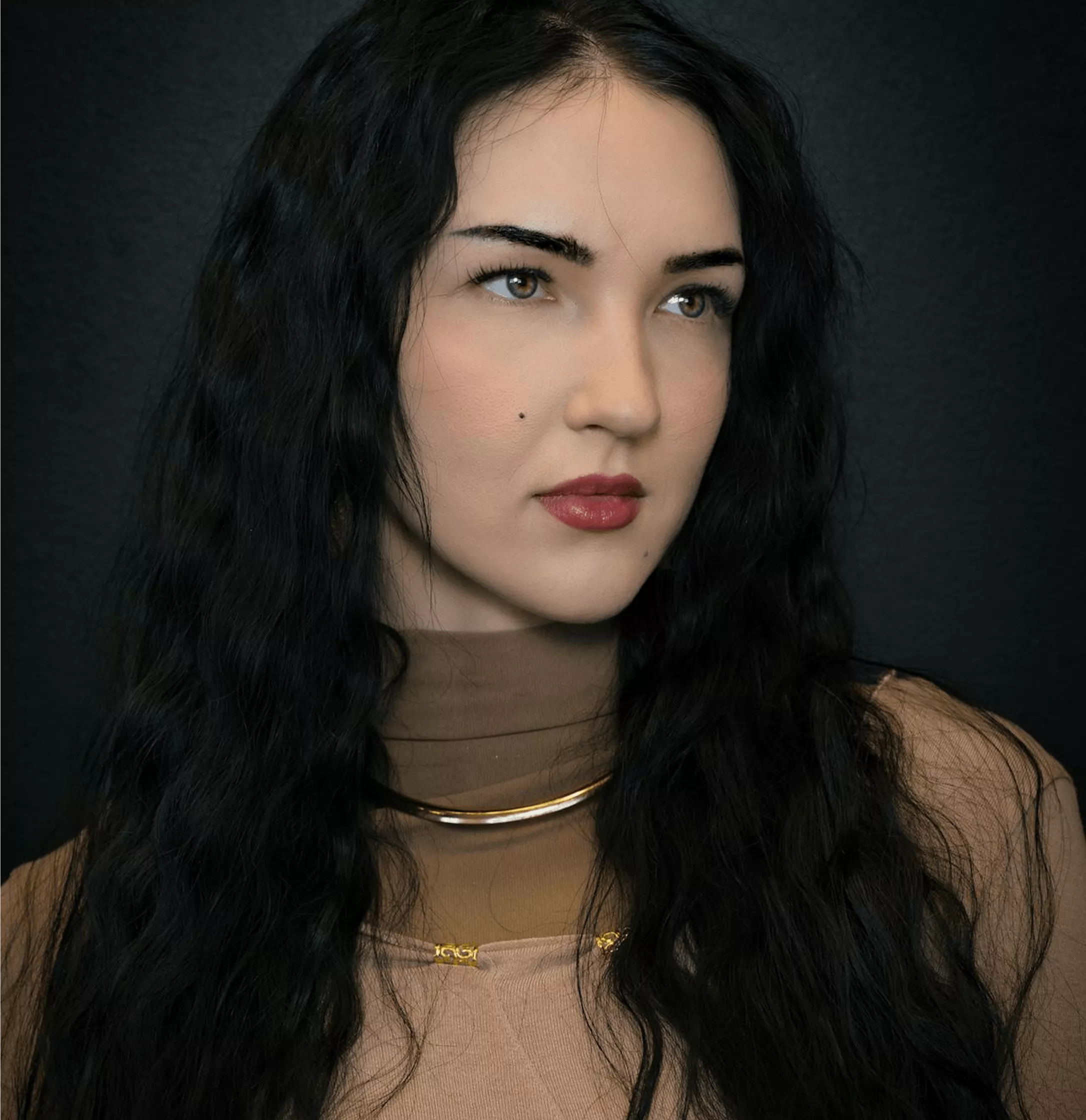 Woman with black wavy mid-length hair wearing sheer tan top with gold collar detail