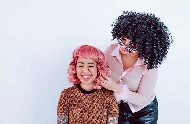 Two people sharing a joyful moment—one with bright pink wavy hair in a patterned orange top, the other with dark curly hair in a pink shirt—celebrating together.
