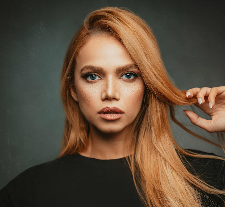 A polished portrait of someone with smooth copper-auburn hair and professional makeup against a muted teal background—conveying expertise and trustworthiness.