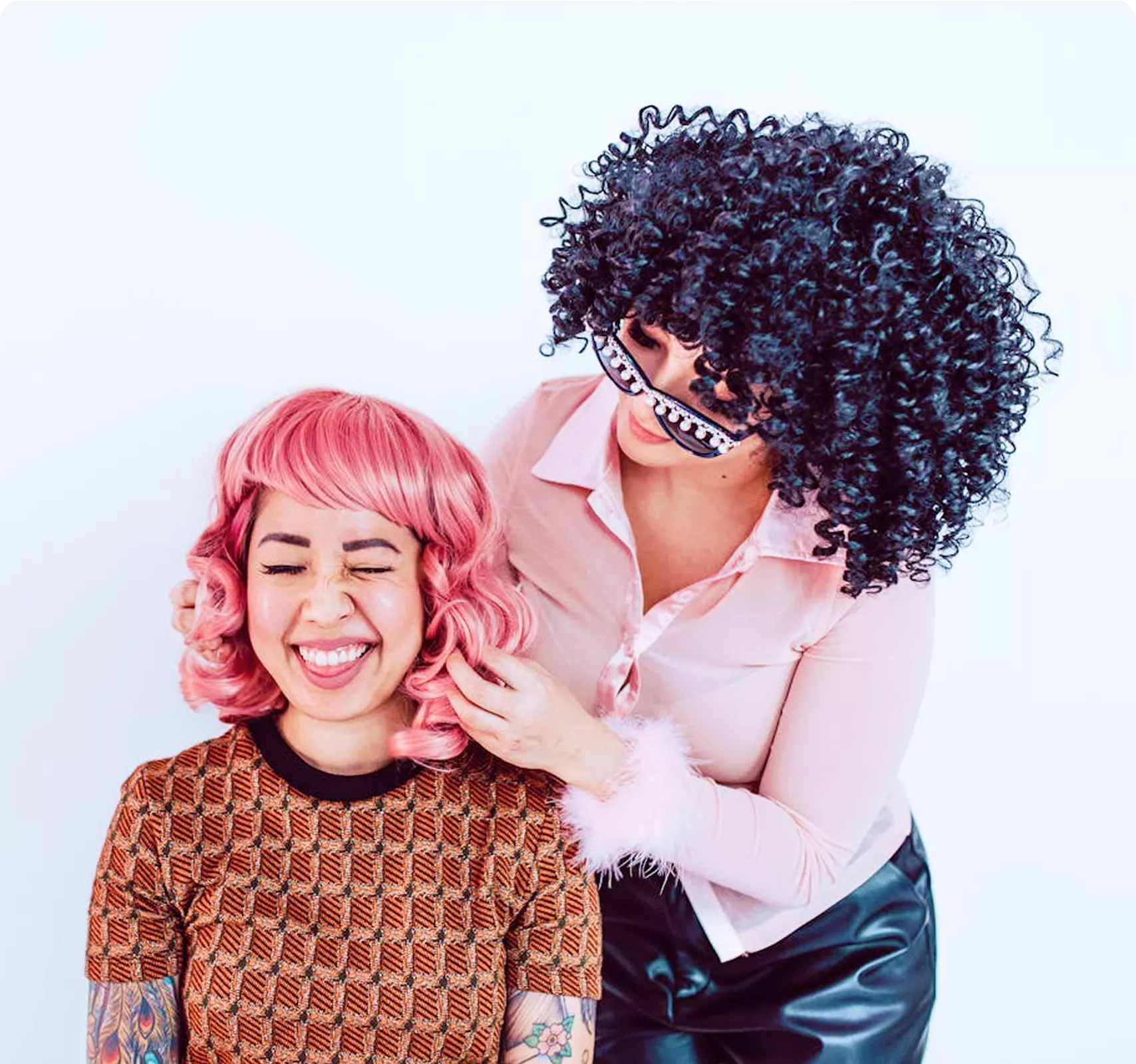 Two people with bold hair colors—one with bright pink wavy hair in an orange patterned sweater, and another with voluminous dark curly hair in a pink shirt—creating a fun, playful composition.