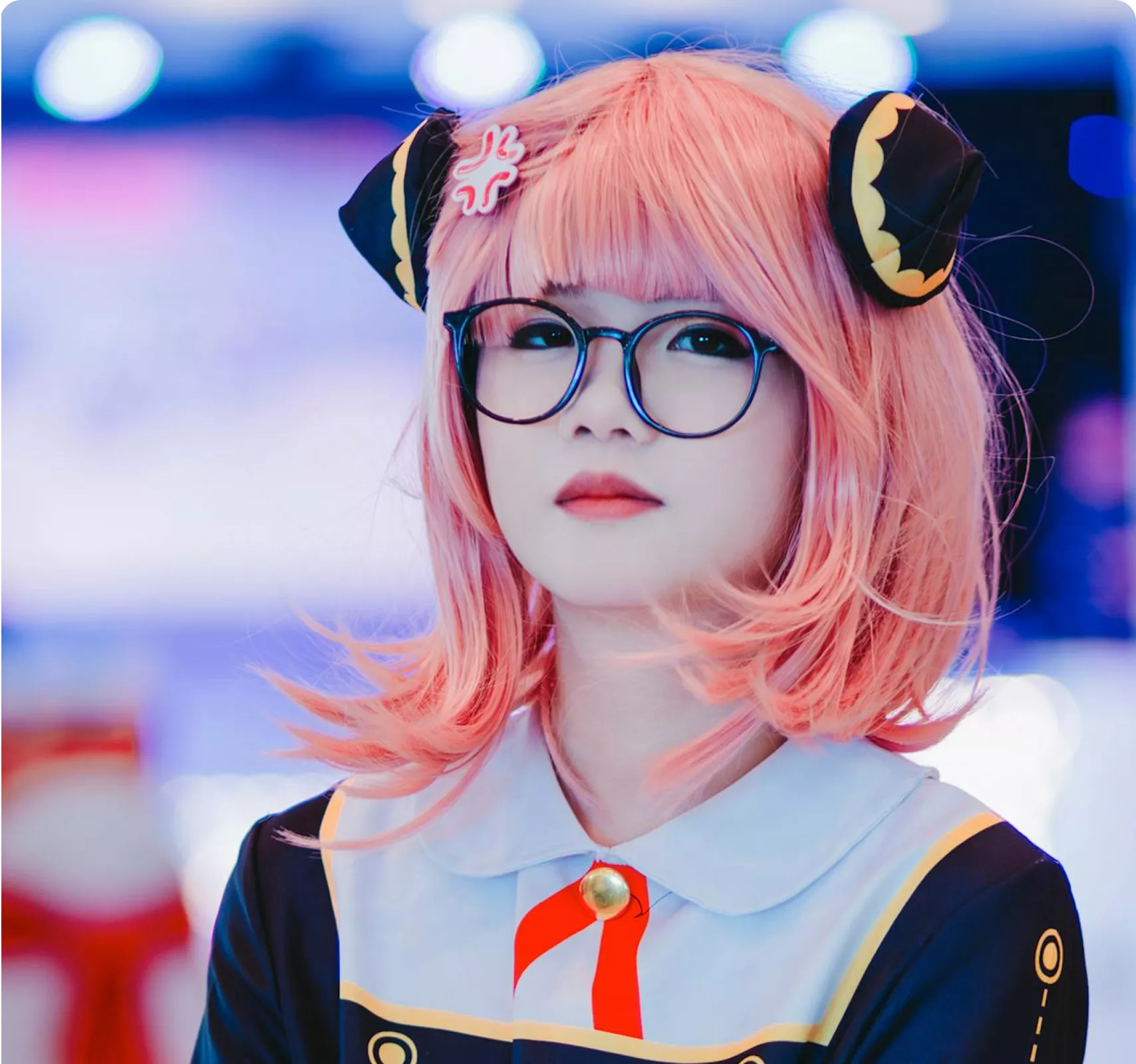 An anime-style character with pastel pink hair, blue round glasses, and black headphones with gold accents, wearing a light blue collar and dark outfit against a dreamy bokeh background.