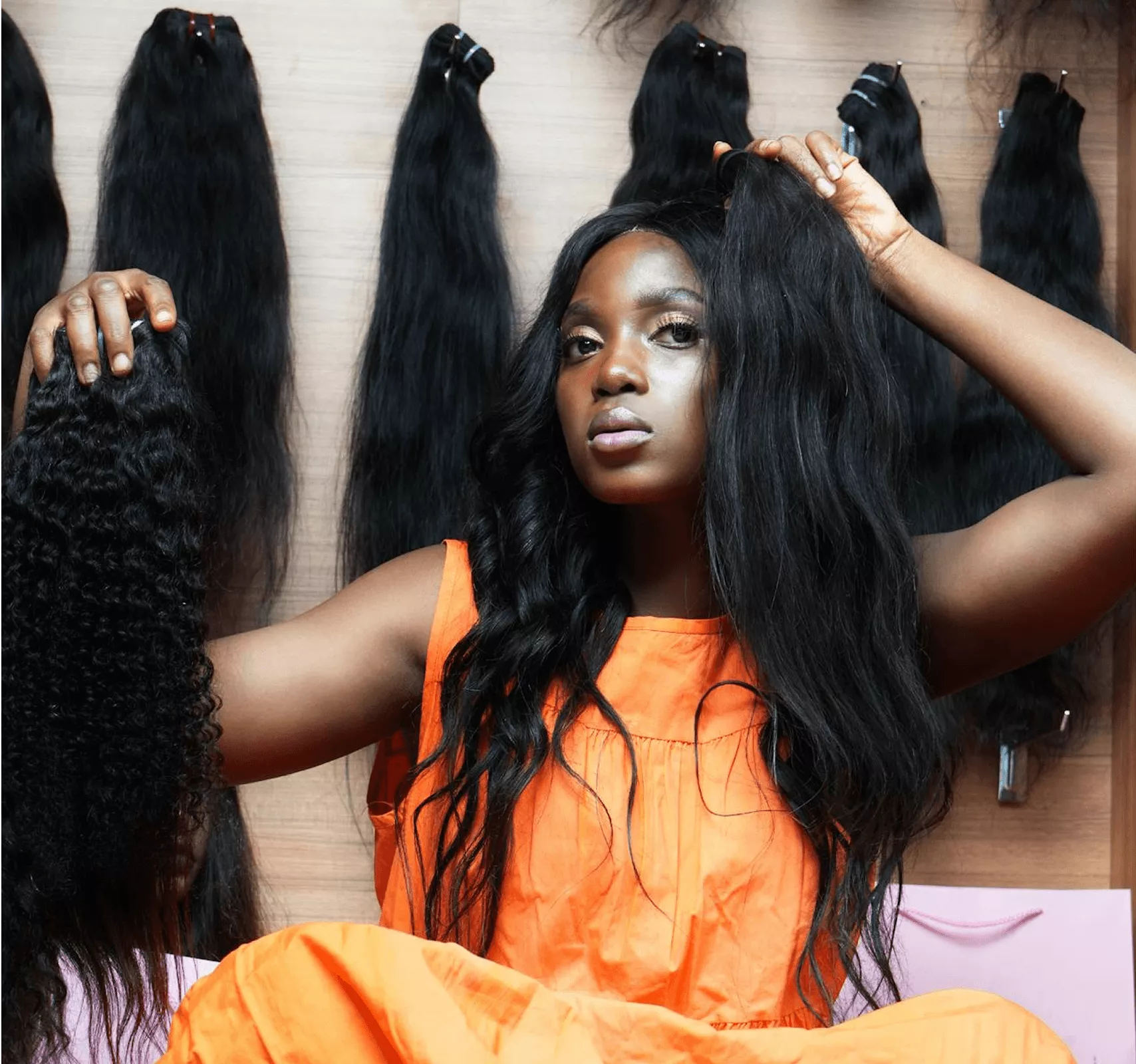 Woman wearing long black wavy wig in orange top posing with display of bundled black wigs background