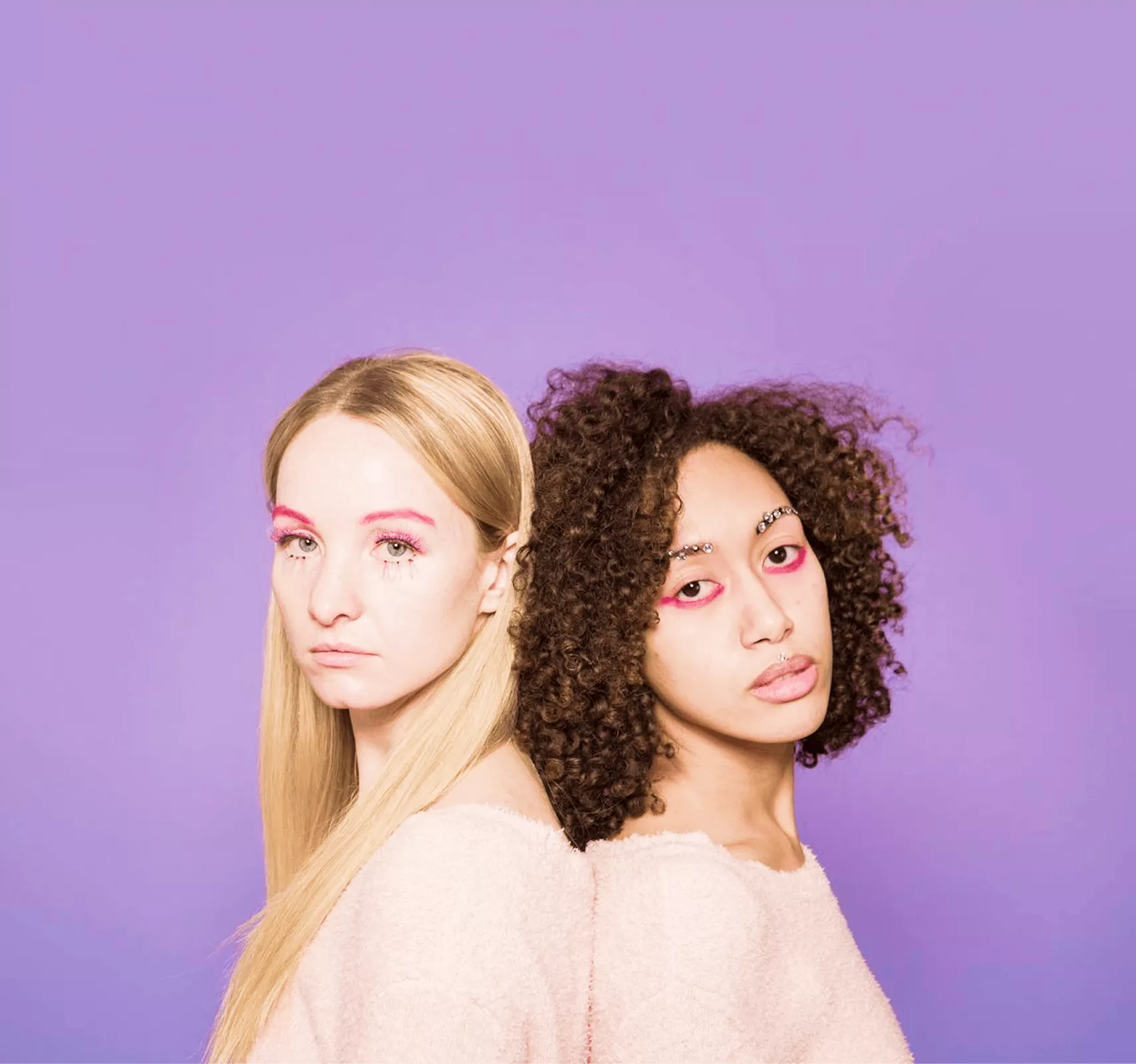Two women back-to-back: blonde straight hair with pink eye makeup and brown curly hair with pink makeup
