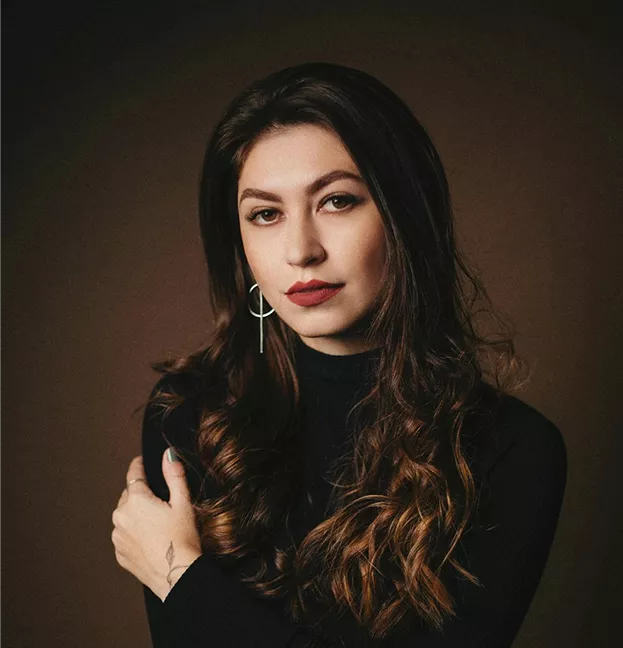 A woman with long wavy dark hair wearing a black turtleneck and hoop earring posing against a warm brown background.