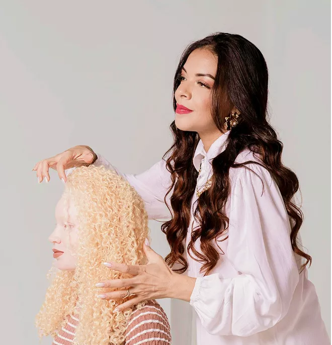 A poised composition showing someone in profile with voluminous blonde curly hair, while another person with long dark wavy hair in a soft pink blouse with a bow detail frames them from behind against a neutral beige background—creating an elegant, artistic portrait.