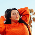 Woman with black wavy mid-length hair wearing orange ribbed knit sweater looking upward outdoors