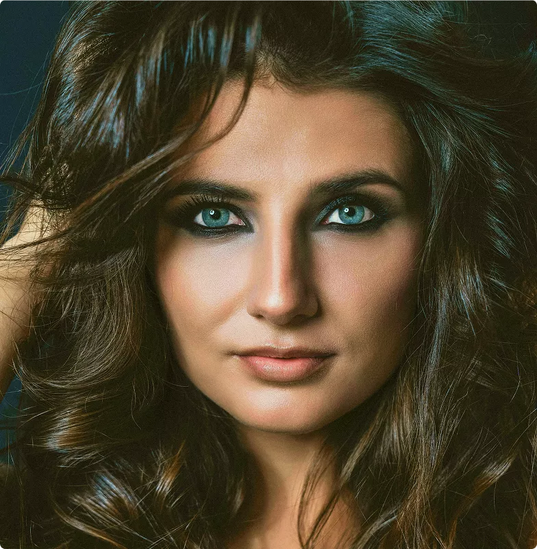 Close-up portrait of a woman with voluminous brown hair and striking blue eyes with smoky eye makeup on a dark background.