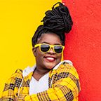 A woman with braided updo hair wearing yellow sunglasses and a yellow plaid jacket against a red and yellow wall.