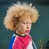 A young person with a voluminous blonde afro wearing a colorful tie-dye top with gold earrings outdoors.